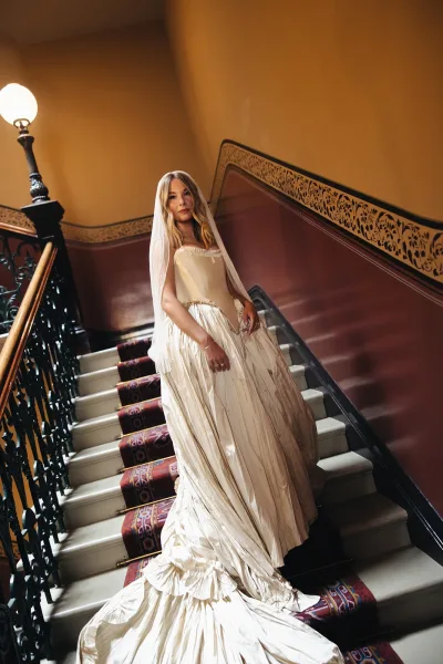 Bridal portrait of a bride on staircase in a satin gown with long veil and ruffled train, framed by an ornate iron banister indoors