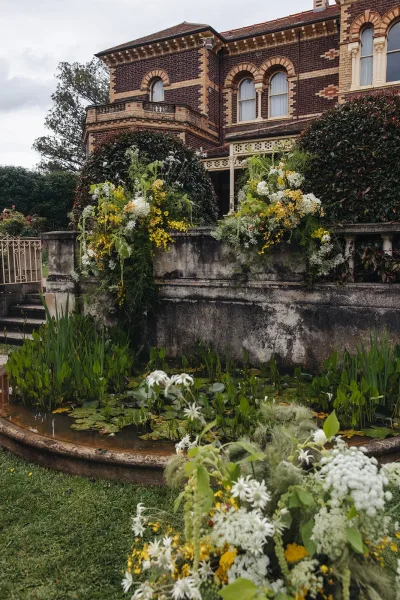 Wedding floral installation with cascading yellow and white blooms and greenery framing a brick mansion entrance by a pond with lily pads