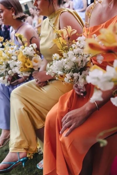 Bridesmaids with bouquets holding yellow and white bouquet mixes of calla lilies, sweet peas, and orchids on an outdoor lawn ceremony