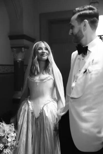 Wedding couple portrait in black and white, bride smiling at groom as they hold hands, veil and bouquet in a hotel hallway by a wall sconce