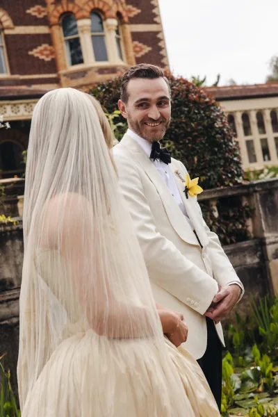 First look moment as groom in a cream tuxedo turns back toward the bride’s long veil outside a brick building with arched windows