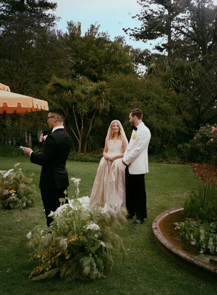 Wedding ceremony moment as bride in veil and strapless dress holds hands with groom in white tuxedo while officiant reads vows in garden