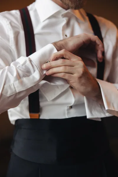 Groom getting ready, buttoning cufflinks on a white dress shirt with suspenders and dress pants in a warm neutral indoor room