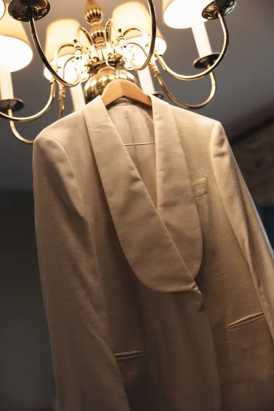 Groom suit detail, wedding suit jacket hanging on a wooden hanger, cream jacket beneath a chandelier against an indoor wall and ceiling