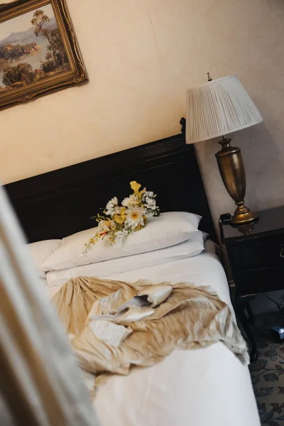 Wedding detail flatlay with bridal shoes on bed beside a yellow and white bouquet, champagne satin robe, and lamp in a classic hotel room