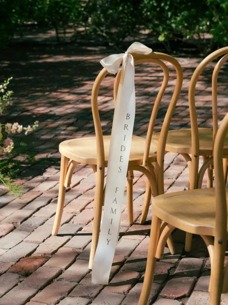 Wedding chair sign reading reserved for family tied with a white ribbon bow on a wooden chair, set on a brick patio with greenery