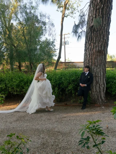 First look moment as bride approaches groom waiting under a tall tree, lifting her dress train and veil along a gravel garden path