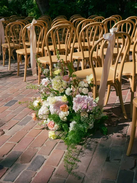 Ceremony aisle decor with outdoor ceremony chairs tied in white ribbons, leading to pastel ground florals on a sunlit brick patio