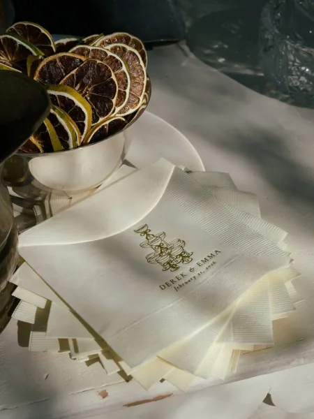 Wedding cocktail napkins with custom wedding napkins monogram in gold foil beside dried citrus slices, cocktail glass, and plate on white bar tablecloth