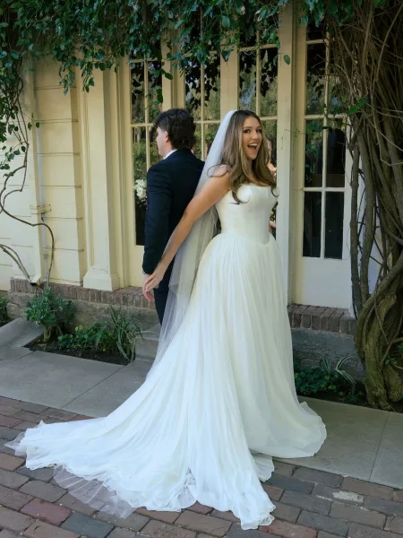 First look moment with bride and groom back to back as she taps his shoulder, her long train and veil on a brick walkway by ivy and French doors