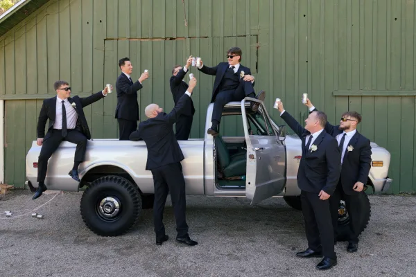 Groomsmen toast with beer cans in black suits and sunglasses, sitting on a vintage pickup truck by a green barn wall