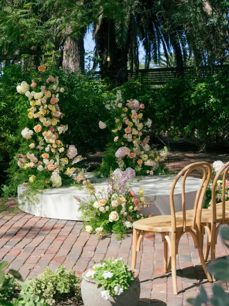 Ceremony floral decor with pastel rose and hydrangea arrangements lining a white curved stage on a brick patio, surrounded by garden shrubs and palms