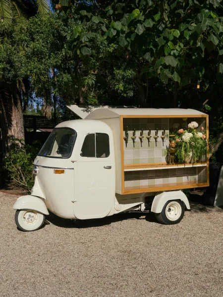 Mobile bar cart with a vintage mobile bar three-wheeler, beer taps, and a greenery garland canopy set on a gravel driveway among tropical trees