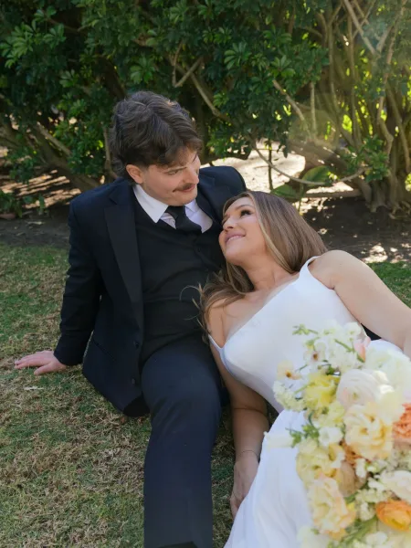 Couple portrait of bride and groom sitting on grass, bride leaning on him holding a pastel bouquet in warm garden sunlight