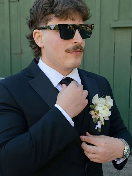 Groom portrait with groom adjusting tie in a black suit, sunglasses, and white rose boutonniere against a green wooden wall