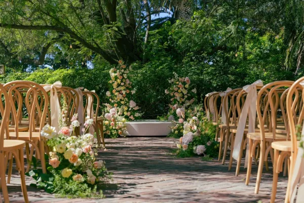 Ceremony aisle decor with floral lined wedding aisle, wood chairs with ribbons framing peach roses and hydrangeas on a sunlit brick path