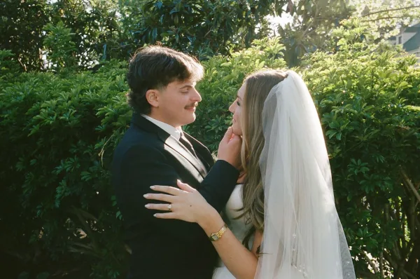 Wedding couple portrait of bride and groom close up embracing as groom holds her face, long veil catching sunlight amid green shrubs and trees