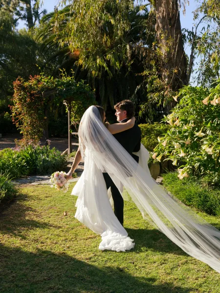 Couple portrait of bride and groom embrace in a garden, her long veil flowing behind as she holds a pastel bouquet near a pergola