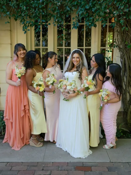 Bridesmaid group photo with bride and bridesmaids holding bridesmaid bouquets in pastel dresses by ivy-covered windows on a brick walkway
