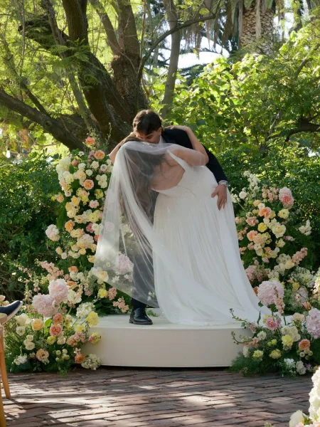 Wedding kiss as groom dips bride under her veil on a raised garden ceremony stage, framed by pastel roses, hydrangeas, and greenery in sunlight
