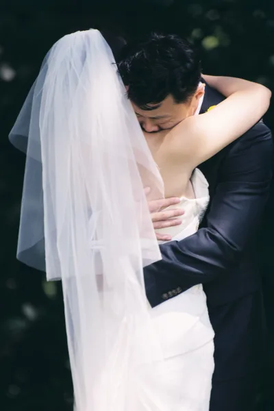 Wedding hug as bride and groom embrace, her long veil draped over a strapless dress while he wears a suit with boutonniere, outdoors amid blurred greenery
