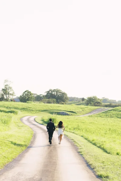 Couple portrait of bride and groom walking away hand in hand on a country road through green fields and rolling hills at sunset