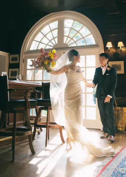 Wedding couple portrait of bride and groom holding hands, bride twirling in satin gown and veil with bouquet by arched window at bar