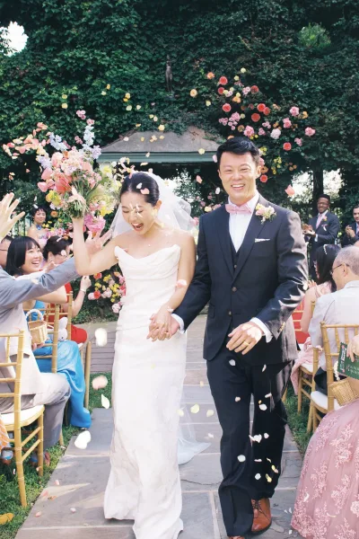 Wedding recessional newlyweds walking down aisle holding hands as guests toss petals under a floral arch in a garden, bride raising bouquet