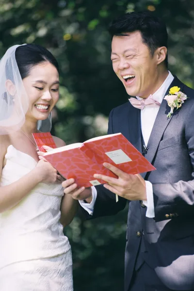Ceremony moment of bride and groom laughing as they read wedding vows from a red booklet, her veil and his tuxedo set against trees
