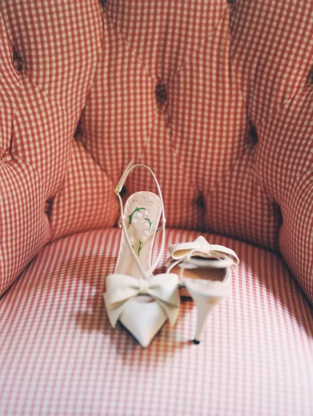 Wedding shoes with white bow heels and ankle strap sandals, featuring an illustrated insole on a pink gingham tufted chair