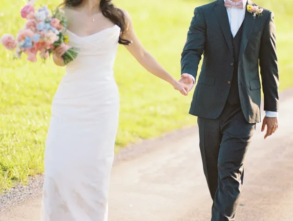 Couple walking hand in hand, bride and groom holding hands as she carries a pastel bouquet on a sunlit outdoor path with grass