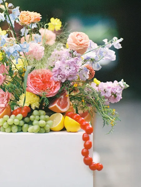 Wedding floral arrangement fruit and flower centerpiece with pastel blooms, grapes, cherry tomatoes, and citrus slices on a white pedestal outdoors