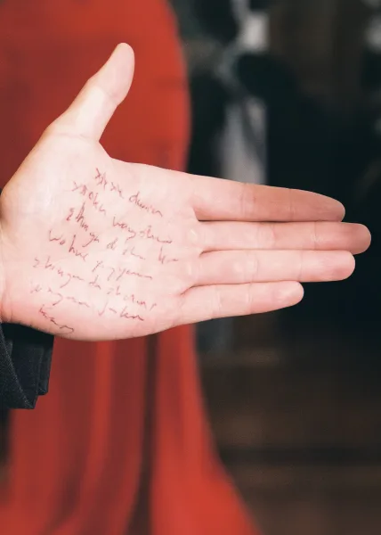 Groom vows notes handwritten on his hand in pen ink, suit jacket cuff visible against a blurred interior with a red wall
