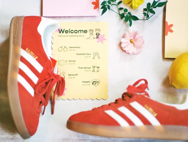 Wedding timeline card with illustrated icons and scalloped edge, styled with red sneakers, paper flowers, and greenery on linen backdrop
