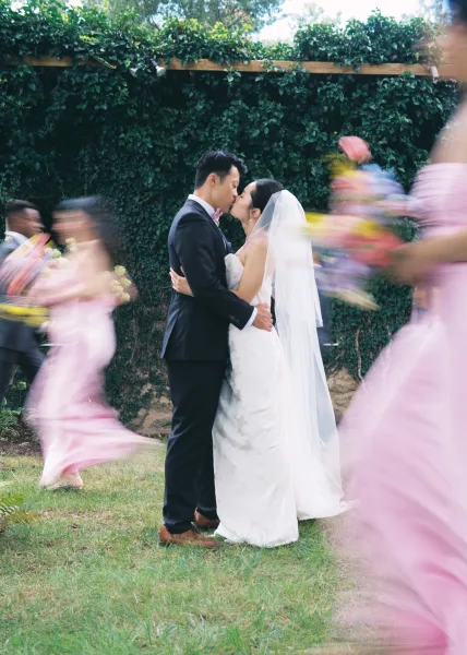 Wedding kiss portrait of bride and groom kissing outdoors, her veil trailing as bridesmaids with bouquets blur past by an ivy wall