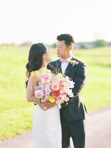 Couple portrait of bride and groom embrace in a green field, bride holding a pastel bouquet, groom in tuxedo with bow tie under open sky