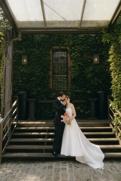 Couple portrait capturing a wedding kiss moment against an ivy wall beside wooden steps on cobblestone, bride cradling a colorful bouquet.
