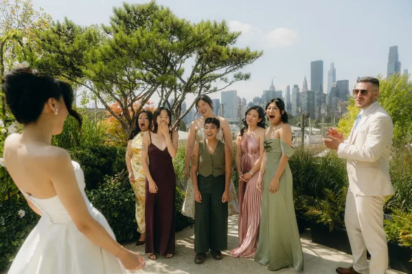 Wedding party first look captures a joyful bride reveal moment, bridesmaids in mismatched dresses on a rooftop garden against a city skyline.