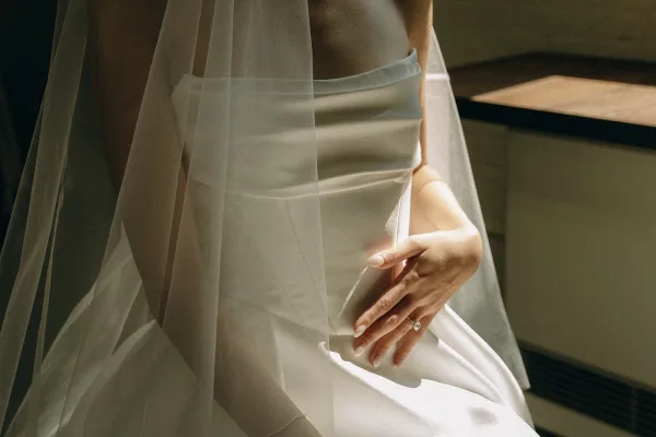 Wedding dress detail of a strapless wedding dress in satin, with the bride’s hand grazing the structured bodice as a veil brushes her shoulder.