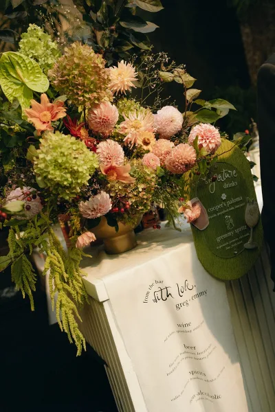Wedding bar decor with a signature drink sign beside lush dahlias, set against dark garden greenery.
