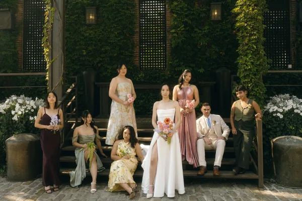 Wedding party portrait with the bride and bridesmaids gathered on wooden steps against an ivy-covered wall, holding bouquets.