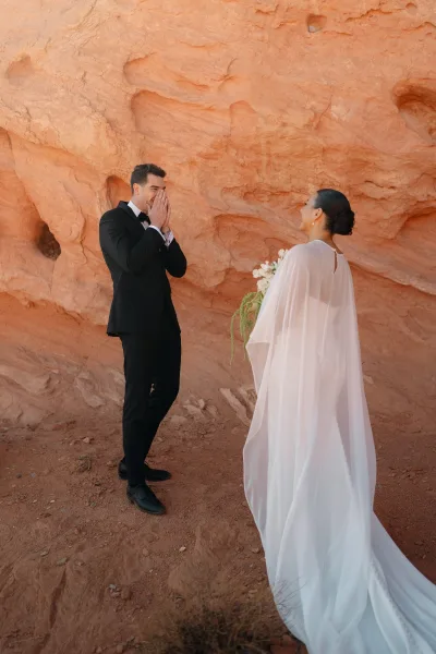 First look moment as bride approaches groom with a simple bouquet, cape veil, and black tux, set against a red rock desert cliff