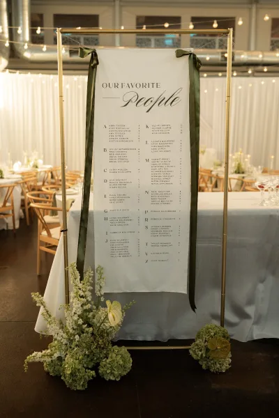 Wedding seating chart fabric seating chart banner on a gold frame with green ribbons and floral base, set amid string-lit reception tables and cross back chairs