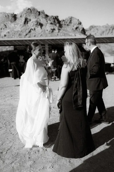 Bride greeting guests, holding a ribbon-wrapped bouquet, laughing with bridesmaid at an outdoor desert pavilion with mountains behind