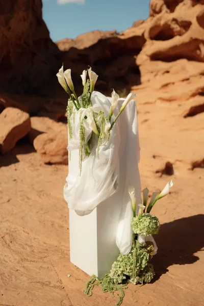 Ceremony aisle marker with calla lilies and greenery on a rectangular pedestal, draped in white fabric against red rock desert sky