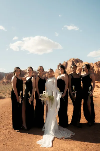 Bridesmaid group portrait of bride with bridesmaids in black dresses holding white calla lily bouquets against red rock desert mountains