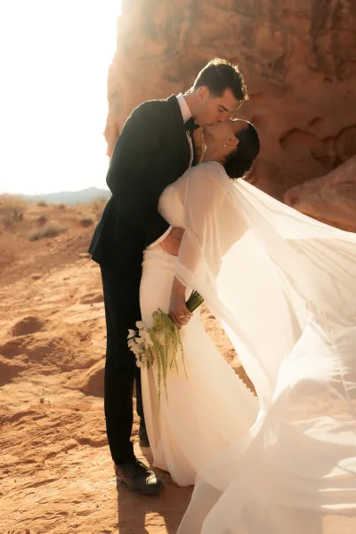 Wedding kiss portrait of bride and groom kiss as her long veil blows in sunlight against red rock desert cliffs and distant hills