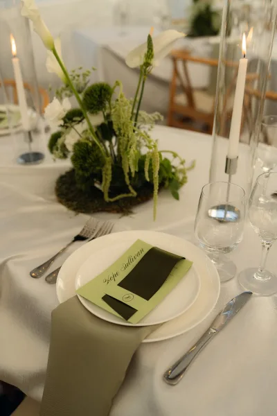 Reception tablescape with wedding place setting, calla lily floral centerpiece, green napkin, menu card, and taper candles in glass hurricanes.