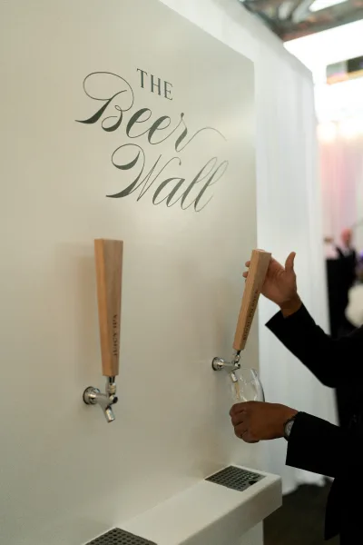 Wedding beer wall with beer taps and wooden handles beneath a custom beer wall sign, featuring a glass and drip tray in an indoor venue