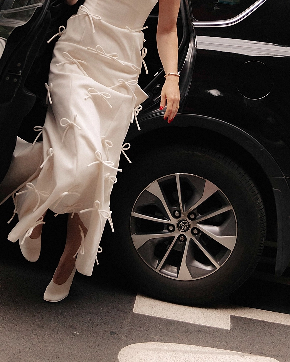 Bridal gown detail with wedding dress bow details in satin fabric, red manicure and bracelet as bride steps from a black car in heels